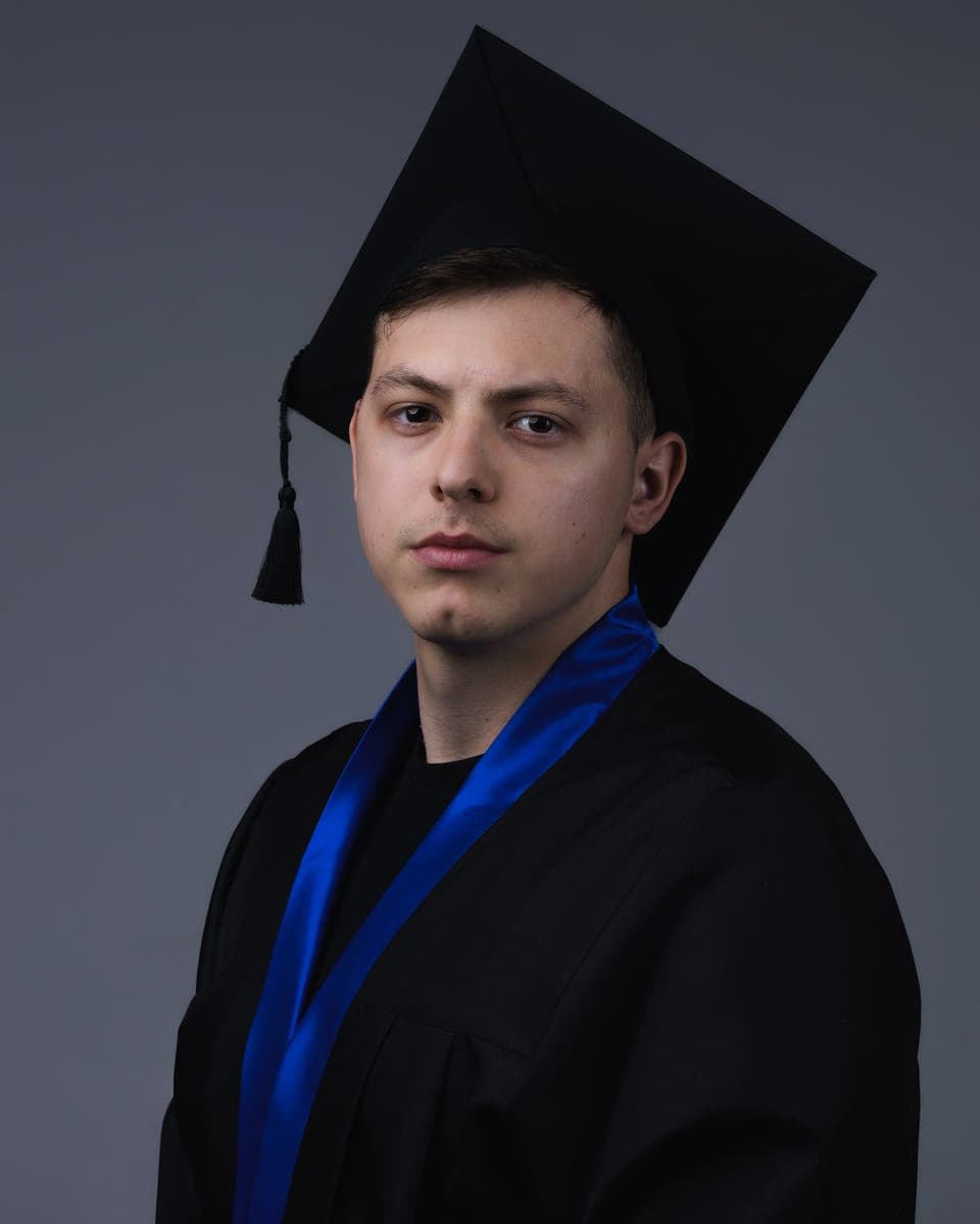 Tarik in a black graduation gown and cap with a royal-blue sash, against a grey studio backdrop