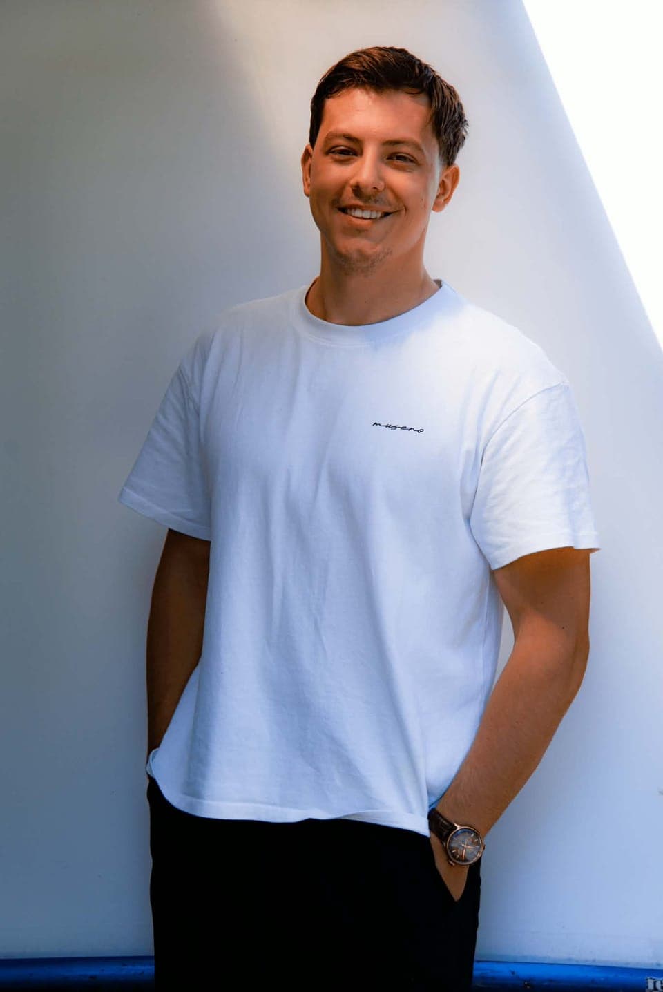 Tarik smiling, hands in pockets, in a white t-shirt against a white wall lit by a slanted beam of sunlight